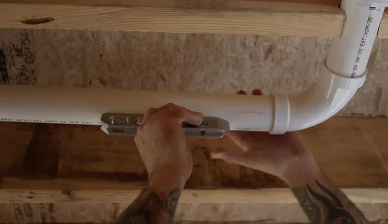 Plumber measuring a white PVC drain pipe between floor joists